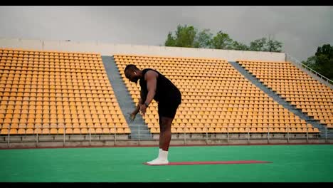 Hombre-Negro-Con-Ropa-Deportiva-Negra-Haciendo-Yoga-En-Un-Estadio-Verde-Con-Sillas-Amarillas-En-Las-Gradas