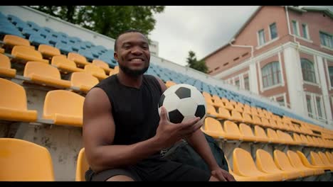 Un-Atleta-De-Piel-Negra-Con-Uniforme-Deportivo-Negro-De-Verano-Posa-En-Las-Gradas-Amarillas-Del-Estadio-Con-Un-Balón-De-Fútbol-En-Las-Manos