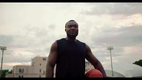 Un-Atleta-Masculino-Serio-Con-Piel-Negra-Y-Una-Camiseta-Negra-Posando-En-El-Estadio-De-La-Ciudad-Con-Una-Pelota-De-Baloncesto-En-Las-Manos-Contra-Un-Cielo-Gris