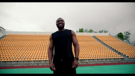 A-Black-man-in-a-black-sports-uniform-poses-and-uses-a-battle-cry-in-front-of-a-stadium-with-yellow-chairs-in-the-stands