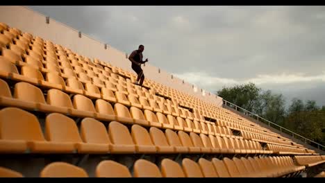 Un-Atleta-De-Piel-Negra-Con-Uniforme-Deportivo-Negro-De-Verano-Y-Auriculares-Bluetooth-Blancos-Camina-Por-El-Estadio-Con-Sillas-Amarillas-En-Las-Gradas