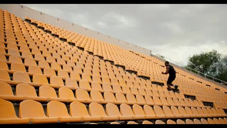 Ein-Sportler-Mit-Schwarzer-Hautfarbe-In-Schwarzer-Sport-Sommerkleidung-Steigt-Die-Treppe-Im-Stadion-Hinauf,-Mit-Gelben-Stühlen-Auf-Der-Tribüne.-Sport-Im-Stadion