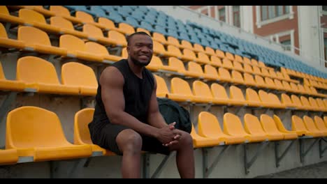 Un-Atleta-De-Piel-Negra-Con-Uniforme-Deportivo-Negro-De-Verano-Se-Sienta-En-Las-Gradas-Del-Estadio,-Mira-La-Cámara-Y-Sonríe