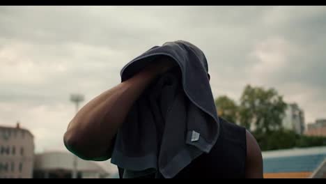 A-black-skinned-sportsman-in-a-black-jersey-wipes-his-face-and-drinks-water-from-a-special-bottle-at-a-city-stadium-against-a-gray-sky