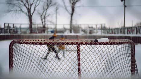 Dos-Jóvenes-Niños-Canadienses-Vestidos-Con-Ropa-De-Invierno-Juegan-Hockey-En-Una-Pista-De-Hockey-Al-Aire-Libre-En-Una-Pequeña-Comunidad-Rural