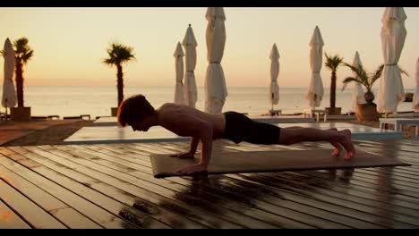 Ein-Mann-Mit-Nacktem-Oberkörper-Macht-Morgens-Yoga.-Trainiert-Die-Katze-Auf-Einer-Speziellen-Matte-Am-Strand,-Die-Mit-Brettern-Bedeckt-Ist