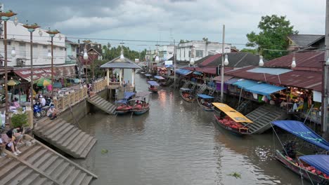 Ein-Boot,-Das-Vom-Fluss-Mae-Klong-In-Den-Schwimmenden-Marktkanal-Amphawa-Einfährt-Und-An-Reihen-Von-Souvenirläden,-Restaurants-Und-Geparkten-Holzbooten-Am-Kanalufer-In-Samut-Songkhram,-Thailand,-Vorbeifährt