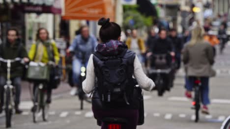 Mujeres-Jóvenes-Subiéndose-A-Su-Bicicleta-Y-Recorriendo-Una-Calle-Muy-Transitada-En-Ámsterdam,-Países-Bajos.