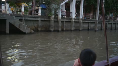 Cruising-along-the-banks-of-Amphawa-Floating-Market-canal,-a-little-boy-is-looking-at-his-surroundings-as-the-boat-is-moving-towards-Mae-Klong-River-in-Samut-Songkhram-province-in-Thailand