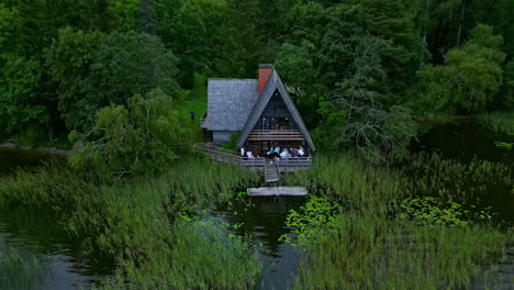 Primer-Plano-De-Una-Toma-De-Un-Dron-De-Una-Casa-En-Un-Lago-Con-Gente-Celebrando-Y-Saludando-A-La-Cámara