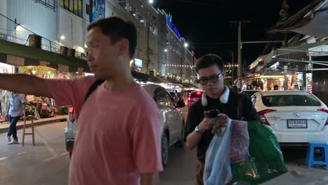 Late-night-shoppers-walking-around-Chatuchak-Weekend-Night-Market,-pointing-to-the-other-shops-on-the-other-side-of-the-road-for-more-wholesale-and-retail-products-to-choose-from,-in-Bangkok,-Thailand