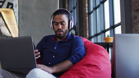 Happy-mixed-race-man-sitting-in-cafe-listening-to-music-on-headphones-using-smartphone-and-laptop