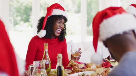 Un-Hombre-Mayor-Afroamericano-Y-Una-Mujer-Joven-Con-Sombreros-De-Papá-Noel-Hablando-Y-Sonriendo-Mientras-Están-Sentados-En-El-Comedor