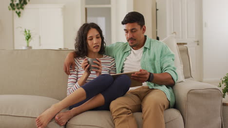 Hispanic-couple-sitting-on-sofa-looking-at-tablet-discussing