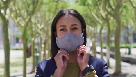 Portrait-of-african-american-woman-putting-on-face-mask-looking-at-camera-in-street