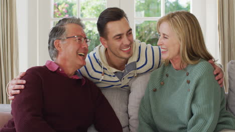 Retrato-En-Video-De-Un-Feliz-Esposo-Y-Esposa-Caucásicos-Sentados-En-El-Sofá-Con-Un-Padre-Mayor-Sonriendo