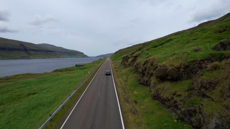 Tracking-aerial-shot-of-car-driving-on-Faroese-road-between-mountain-and-fjords