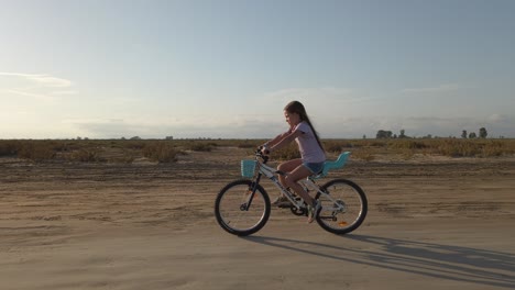 Kleines-Mädchen-Fährt-Mit-Dem-Fahrrad-Auf-Einer-Sandigen-Straße,-Während-Die-Sonne-In-Der-Abenddämmerung-Scheint