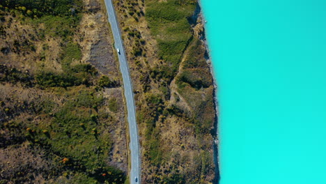Top-down-aerial-drone-view-of-a-car-driving-along-a-scenic-mountain-road-beside-a-turquoise-glacial-lake