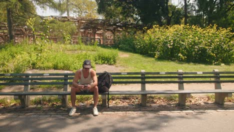 Man-sitting-on-park-bench-checks-phone-looks-up,-standing-around-walks-out-of-empty-frame