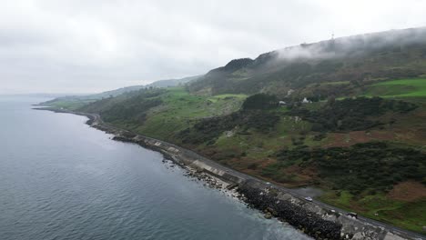 Aerial-panning-shot-over-the-beautiful-rocky-landscape-on-the-coast-of-Northern-Ireland-coastal-road-near-Glenarm-town-with-view-of-the-rocky-road,-the-blue-sea-and-rising-mountain-in-the-morning
