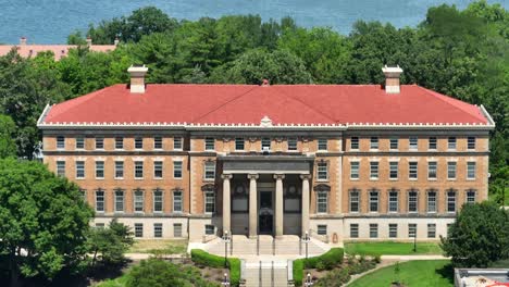 Vista-Aérea-De-Un-Edificio-Histórico-De-Ladrillo-Con-Techo-De-Tejas-Rojas,-Ubicado-Entre-Exuberantes-árboles-Verdes-En-El-Campus-De-La-Universidad-De-Wisconsin.