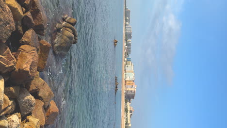 vertical-video-with-copy-space-of-the-Mediterranean-Sea,-with-rocks-in-the-foreground-and-a-city-of-Blanes-in-the-background-on-the-Costa-Brava-of-Girona