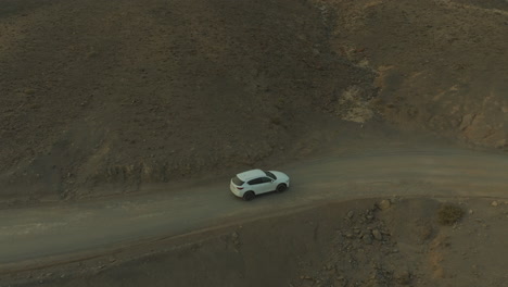 Insel-Fuerteventura:-Fantastische-Luftaufnahme-Mit-Seitlicher-Verfolgung-Eines-Weißen-Autos,-Das-Auf-Einer-Unbefestigten-Straße-In-Einer-Wüsten--Und-Vulkanumgebung-Fährt