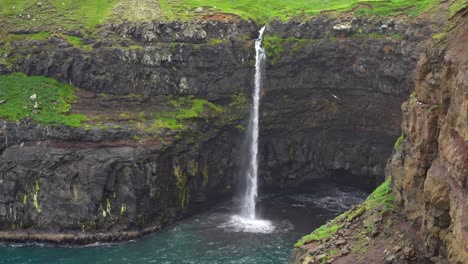 Mulafossur-Waterfall-in-the-Faroe-Islands-with-distant-view-of-person-hiking-on-top-of-cliff