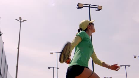 La-Mujer-Juega-Un-Servicio-De-Pádel-Bajo,-Juego-De-Pádel-En-La-Playa,-Deportes-Soleados-Al-Aire-Libre