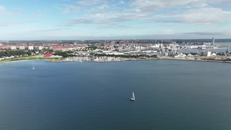 A-sailboat-cruising-the-bay-of-Aarhus-Denmark---Summer-aerial-with-panoramic-cityscape-background