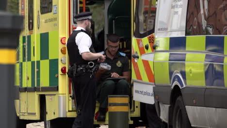 Two-paramedics-talk-with-a-Metropolitan-police-officer-next-to-a-parked-ambulance-and-a-police-van-at-the-scene-of-an-incident