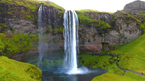 Drohnenaufnahme-Eines-Großen-Wasserfalls-Im-Süden-Islands-Mit-Viel-Grün-Rundherum,-Einem-Sonnenstrahl-Und-Touristen