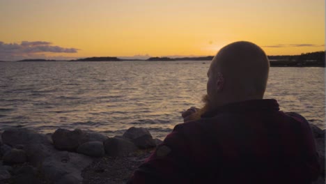adult-Caucasian-male-with-ginger-beard-sitting-by-himself-on-rocky-beach-wearing-red-and-black-shirt-no-hair-looking-over-the-horizon-of-the-vibrant-sunset-thinking-about-life-smoking-antique-pipe