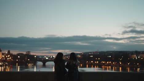 Two-women-on-bridge-gaze-at-illuminated-cityscape-at-dusk-in-Prague