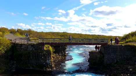 Ein-Mann-Auf-Einer-Brücke,-Der-Seine-Drohne-Steuert,-Die-Sich-Allmählich-Von-Ihm-Entfernt-Und-Den-Blick-Auf-Eine-Herrliche-Isländische-Landschaft-Freigibt:-Den-Blauen-Wasserfall-Von-Brúarfoss