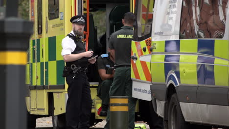 Two-paramedics-talk-with-a-Metropolitan-police-officer-as-he-listens-to-his-radio-next-to-a-parked-ambulance-and-a-police-van-at-the-scene-of-an-incident