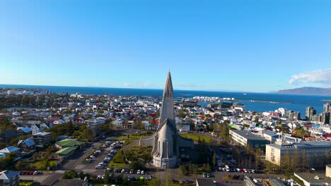 Toma-De-Drone-De-Una-Hermosa-E-Impresionante-Iglesia,-La-Hallgrímskirkja,-En-La-Capital-De-La-Isla-De-Reykjavik,-Con-La-Ciudad-Alrededor,-El-Mar-Al-Fondo-Y-Un-Clima-Soleado.