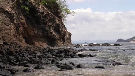Ocean-waves-crashing-on-rocky-shores-of-Playa-Penca-as-fishing-boat-drives-in-background,-Costa-Rica