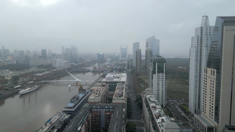 Panoramic-aerial-view-of-highrise-buildings-alongside-the-river-Plate-with-Woman's-Bridge-under-foggy-sky-due-to-climate-change,-Buenos-Aires,-Argentina