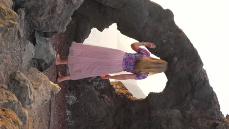 Blonde-woman-explores-scenic-rock-arch-at-sea-cliffs-of-Tenerife-at-golden-hour,-gazes-out-to-sea