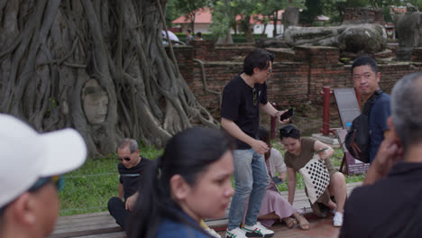 Escena-Cautivadora-De-Personas-Escuchando-Atentamente-Una-Narración-Histórica-Frente-A-La-Cabeza-De-Buda-Enclavada-Entre-Las-Raíces-De-Un-árbol-En-Ayutthaya,-Tailandia.