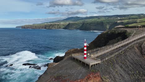 Red-and-white-Fenais-da-Ajuda-lighthouse-on-rugged-rocky-headland,-aerial-dolly