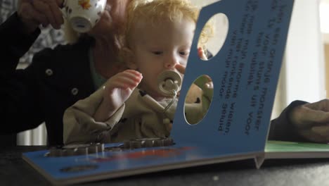 Cute-toddler-girl-turning-pages-of-a-childrens-book-on-top-of-grandmother-her-lap,-while-drinking-coffee-at-the-kitchen-table