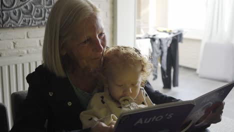Caucasian-Grandmother-reading-a-book-to-her-toddler-grandchild-at-the-kitchen-table