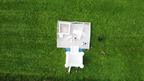 Top-down-aerial-view,-white-bouncy-castle-with-toddlers-playing-in-a-grassfield
