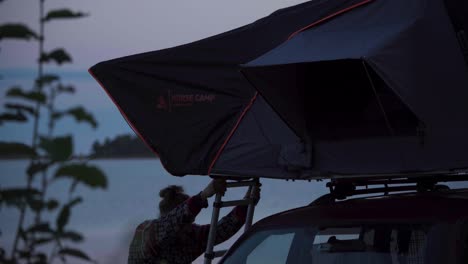 Hombre-Con-Una-Tienda-De-Campaña-En-El-Techo-Del-Coche-Cerca-De-Un-Tranquilo-Lago-Al-Atardecer