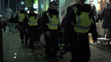 A-unit-of-Metropolitan-riot-police-in-protective-black-overalls-and-Nato-helmets-on-their-belts-march-through-a-crowd-while-policing-a-public-order-incident-at-night