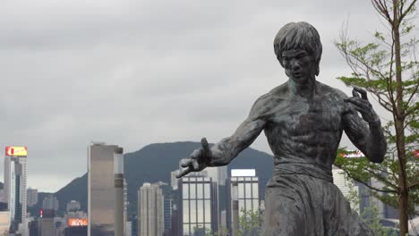 Bruce-Lee-Statue-On-Star-Street-In-Tsim-Sha-Tsui-with-skyscrapers-in-the-background-on-a-busy-day,-Hongkong