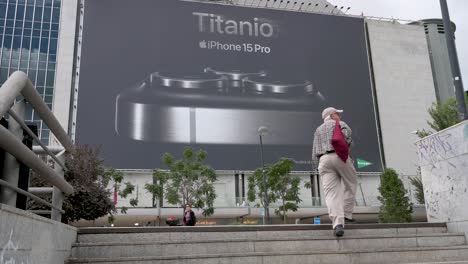 A-large-commercial-billboard-ad-hangs-at-a-shopping-mall-building-announcing-the-Apple-iPhone-15-Pro-for-sale-as-a-shopper-walks-past-the-frame